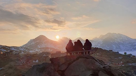 Tre personer sidder på en bjergtop og kigger på solnedgangen. Solen går ned over bjergtoppe som er længere væk. Der ligger sne omkring dem, så de har varmt tøj på.