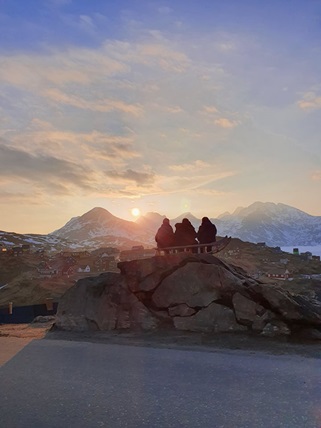 Tre personer sidder på en bjergtop og kigger på solnedgangen. Solen går ned over bjergtoppe som er længere væk. Der ligger sne omkring dem, så de har varmt tøj på.